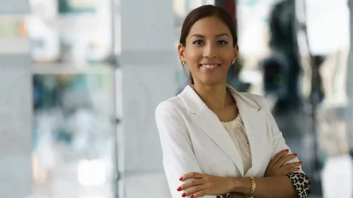 Woman in a white blazer stands with arms crossed, smiling. The image conveys professionalism, confidence, and a welcoming attitude.