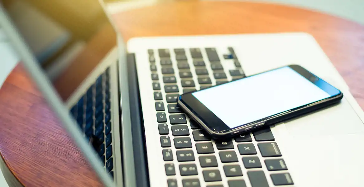 A smartphone rests on the keyboard of an open laptop on a round table. The image symbolizes Alpidus' accessibility and multi-channel communication.
